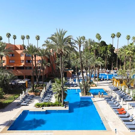 Hotel Kenzi Rose Garden Marrakesch Exterior foto The photo shows a resort-style area with several swimming pools surrounded by palm trees. The pools are clear blue and are lined with numerous lounge chairs for relaxation. There is a vibrant atmosphere, highlighted by lush greenery and a sunny sky. 