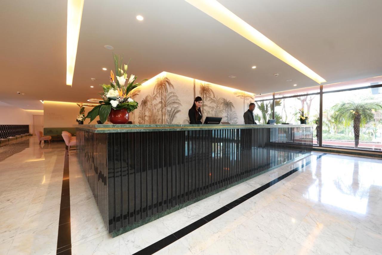 Hotel Kenzi Rose Garden Marrakesch Exterior foto The image shows a modern reception area, likely for a hotel or office building. There is a stylish front desk made of dark materials with a smooth surface. Behind the desk, two staff members are engaged in their work, possibly checking in guests or a